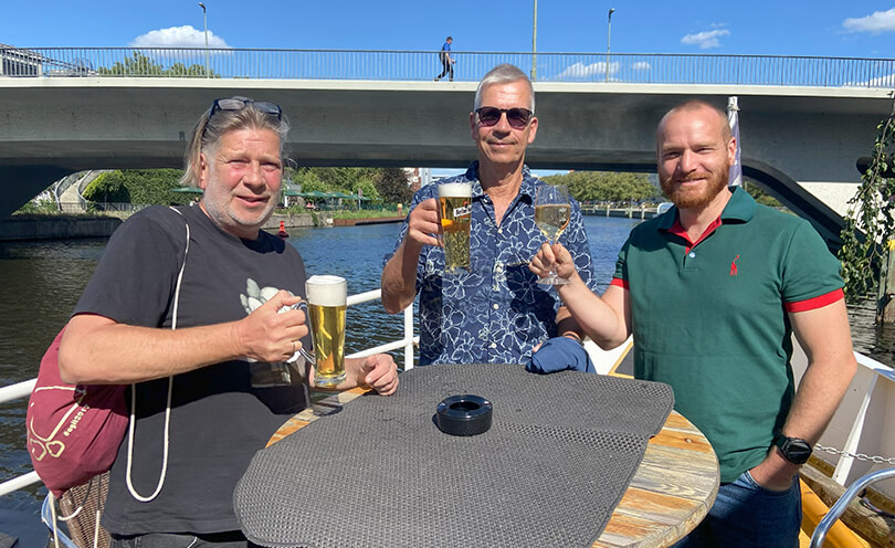 Drei Personen auf einem Boot an einem Tisch, zwei mit Bier eine mit Wein in der Hand. Sie lächeln.