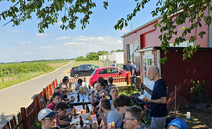 Viele Personen an einem langen Tisch auf einem Hof mit Land drum herum. Sie essen und trinken im Schatten.