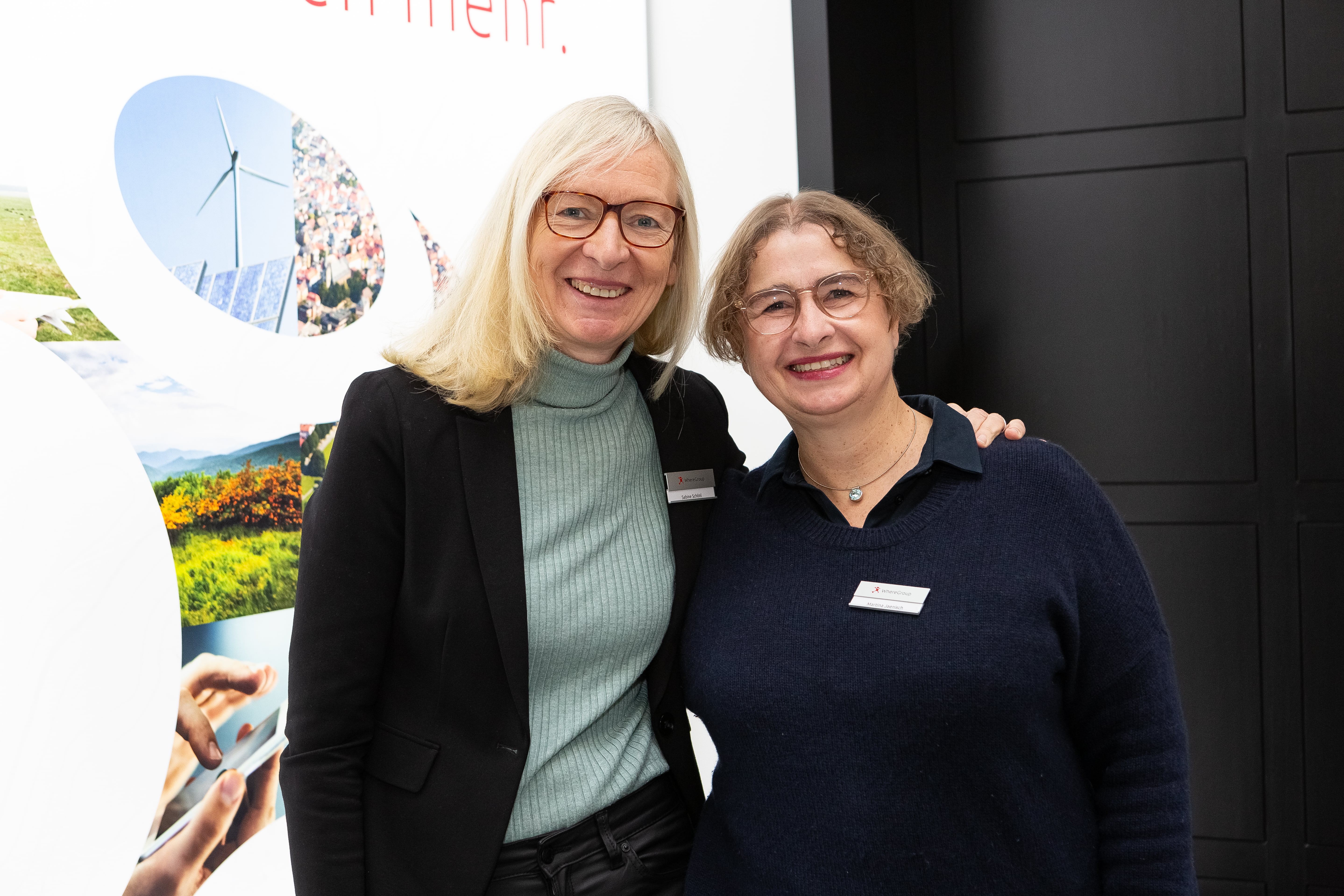 Zwei Damen vor einem schwarz-weißen Hintergrund (Where2B). Sie sind etwas schicker angezogen und tragen Namensschilder. Sie lächeln.